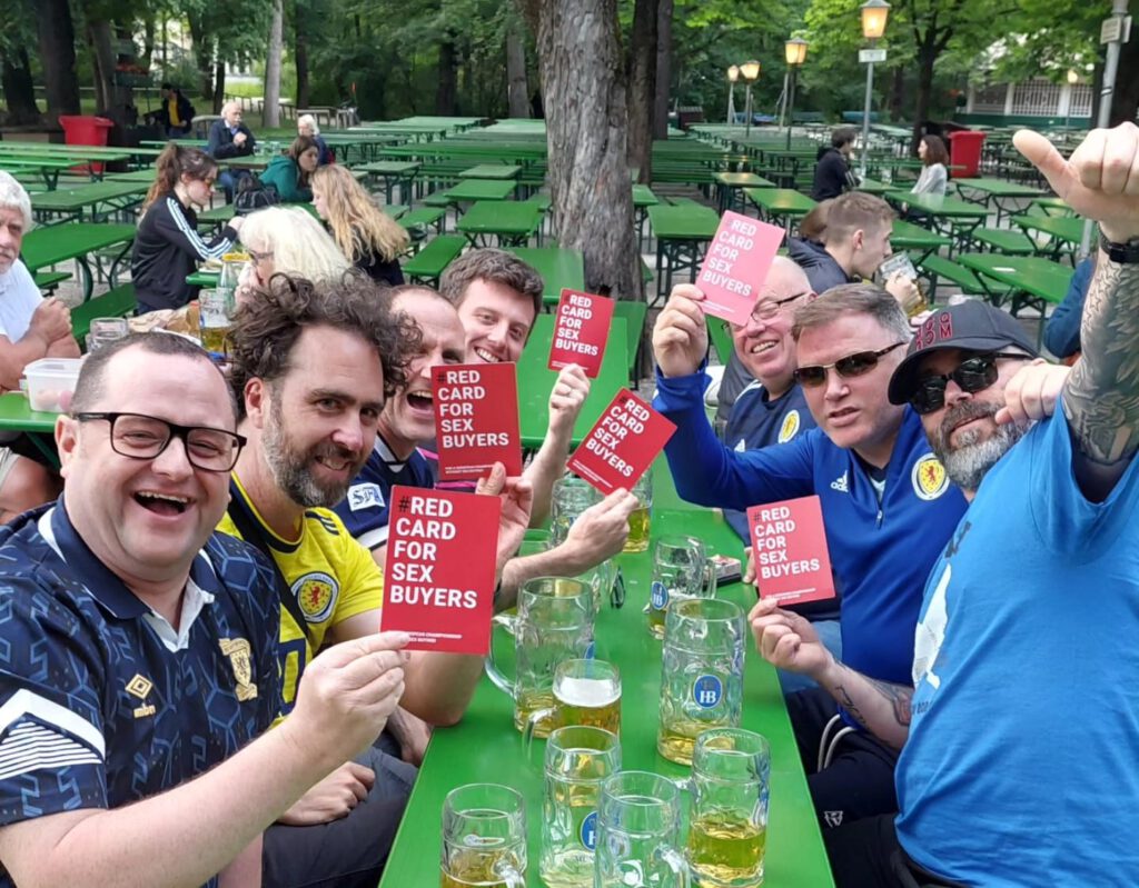 Hier ist Bild 1 zu sehen. Es zeigt eine Gruppe Fußballfans im Biergarten am Chinesischen Turm in München mit den roten Karten der Kampagne.