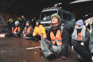 Hier ist das Titelbild zu sehen. Es zeigt Judith Schäfer im November 2023, auf die Straße geklebt, bei einer Blockadeaktion der Letzten Generation auf der Puschkinallee in Berlin.