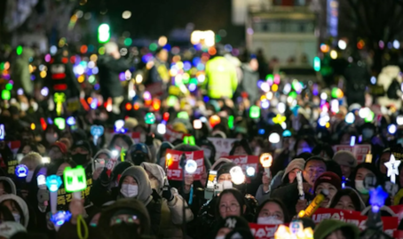 Hier ist das Titelbild zu sehen. Es zeigt eine Demonstration mit vielen Handy-Lichtern in Seoul im Dezember 2024.