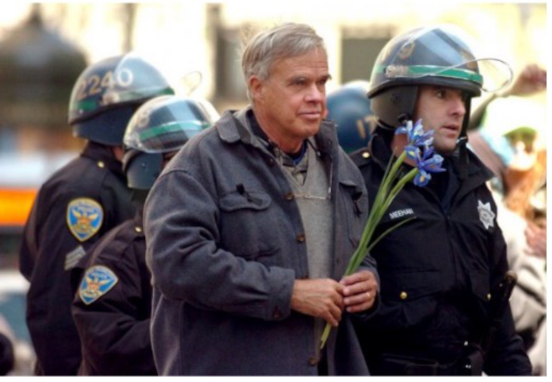 David Hartsough umringt von drei Polizisten, die ihn festnehmen. Er selbst hat Blumen in der Hand.