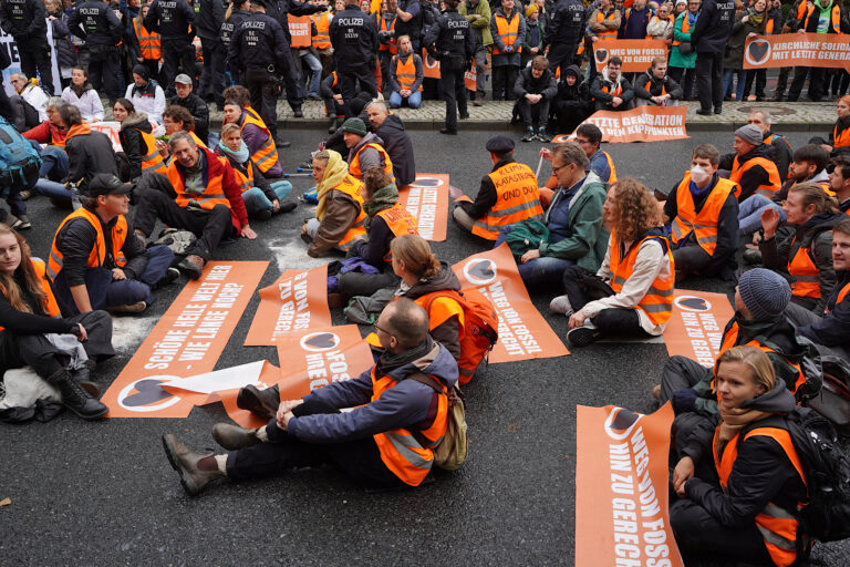 Hier ist das Titelbild zu sehen. Es zeigt eine Protestaktion der Letzten Generation auf der Straße des 17. Juni in Berlin Tiergarten.