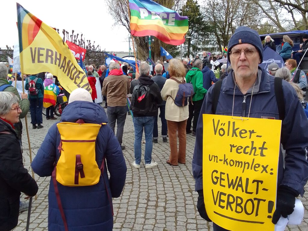 Hier ist das Titelbild zu sehen. Es zeigt Armin Lauven bei einer Friedensdemonstration. Er trägt ein Plakat mit der Aufschrift: „Völkerrecht un-komplex: Gewaltverbot!“